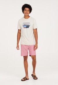 Kortærmet beige t-shirt med blåt og rødt design, kombineret med lyserøde shorts og brune flip-flops. Modell står mod en ensfarvet baggrund.