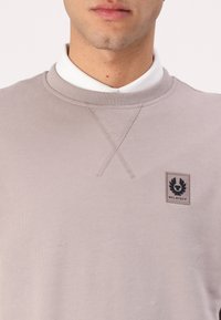 Világosszürke pulóver, texturált felülettel, bordázott gallérral és egy kis fekete Belstaff logóval a bal mellen.