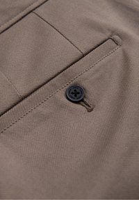 Gros plan sur un tissu beige avec un bouton noir cousu près d'une poche passepoilée oblique sur un vêtement.