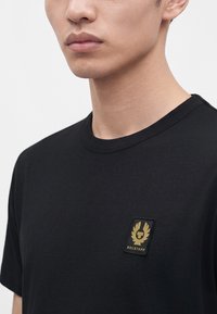 Νέος άνδρας που φοράει απλό μαύρο t-shirt με ένα μικρό χρυσό λογότυπο Belstaff στην αριστερή πλευρά του στήθους, κομμένο στο ύψος των ώμων και του στόματος.
