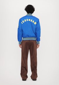 Chaqueta bomber azul con texto amarillo "ICECREAM", puños y dobladillo acanalados, combinada con pantalones marrones, vista desde atrás.