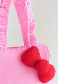 Sac à main rose avec des poignées froncées et un grand nœud en tissu rouge attaché dans le coin supérieur sur fond uni.