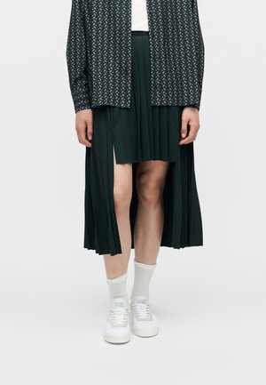 Parte inferiore del corpo di una persona che indossa una gonna plissettata verde scuro con orlo asimmetrico, top a maniche lunghe a motivo, calzini bianchi e scarpe da ginnastica bianche.