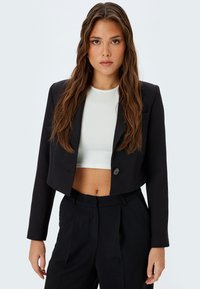 Taillierte schwarze Blazerjacke mit einem einzelnen Knopf, feinem Schnitt und glatter Textur, kombiniert mit einem taillierten weißen Oberteil, was einen formellen Look erzeugt.
