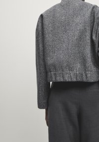 Persoon in een grijs textuurjack met korte mouwen en donkergrijze broek, weergegeven vanaf halverwege de rug tegen een effen lichte achtergrond.
