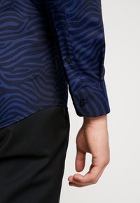 Blå mönstrad skjorta med en vågig design, knäppt manschett och en slät textur. Bärs med svarta byxor, med fokus på skjortans ärmdetail.