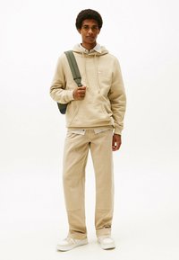 Beige Kapuzenpullover mit Tasche und Kordelzug, über einem gestreiften Shirt getragen. Beige Hosen mit Seitennaht. Kombination mit weißen Sneakers und einem grünen Rucksack.