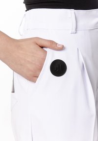 Białe spodnie mają boczną kieszeń z czarnym, okrągłym, skórzanym akcentem. Tkanina wydaje się gładka z delikatnym połyskiem.