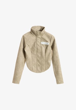 Chaqueta beige de manga larga con cuello alto, costura frontal asimétrica y pequeña etiqueta blanca rectangular en el pecho.