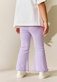 Fialové rebrované zvonové nohavice so zubatými lemami, doplnené bielymi teniskami, pohľad zo zadnej strany.