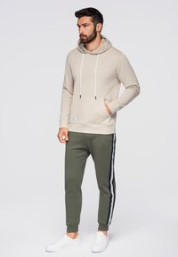 Béžová mikina s kapucí a bílými šňůrkami, s přední kapsou, spárovaná se zelenými tepláky s námořnickými a bílými pruhy po stranách. Bílé tenisky.