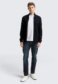 Marine gebreid vest met een rits aan de voorkant en een textuurpatroon, gedragen over een wit t-shirt, gecombineerd met donkere jeans en witte sneakers.