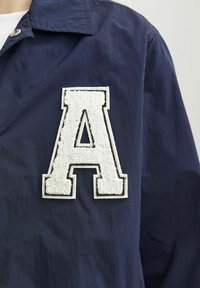 Granatowa kurtka z teksturowaną, haftowaną białą literą "A" na lewej piersi, z klasycznym kołnierzem i gładkim wykończeniem tkaniny.