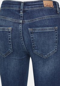 Jeans de ganga azul escuro com bolsos traseiros, costura em contraste e um patch de couro com a marca na cintura. Corte regular, design clássico.