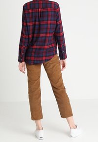 Person wearing a red and navy plaid shirt, brown corduroy pants, and white sneakers, standing facing away on a plain background.