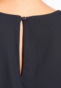 Blusa blu navy con apertura a occhio di serratura chiusa da un bottone nero, caratterizzata da una superficie liscia e un design semplice e pulito.