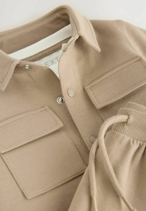 Beige Langarmoberteil mit Kragen, Druckknöpfen, zwei Brusttaschen und elastischen Kordelzugmanschetten. Weiche, glatte Textur.