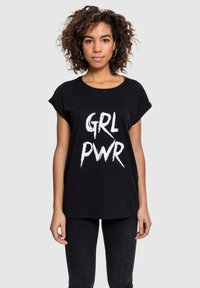 Sort bomulds T-shirt med rulleærmer, der har hvid tekst "GRL PWR" i en fed, distressed skrifttype. Båret med mørke jeans.