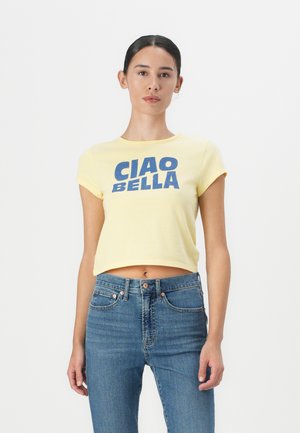 T-shirt cropped jaune avec le texte graphique bleu "CIAO BELLA", col rond et manches courtes. Associé à un jean bleu taille haute.