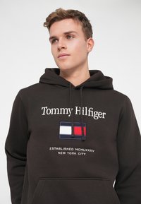 Felpa con cappuccio marrone, con testo ricamato bianco, caratterizzata da un design della bandiera in rosso, bianco e blu. Include una tasca a marsupio frontale.