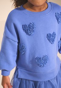 Kék pulóver plüss szív applikációkkal, bordázott nyakkal és mandzsettákkal, laza szabású, világoskék tüll szoknyával párosítva.