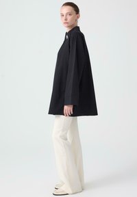 Black oversized shirt with long sleeves, high collar, and a button detail. Paired with cream flared pants and light-colored footwear.