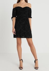 Vestido negro sin mangas con textura floral, mangas cortas, cuerpo ajustado y dobladillo recto. Combinado con tacones negros de tiras.