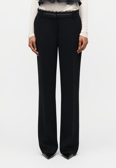 Pantalon noir à jambes larges avec taille ornée de dentelle, porté avec des chaussures pointues noires et un haut blanc à manches longues sur un fond uni.