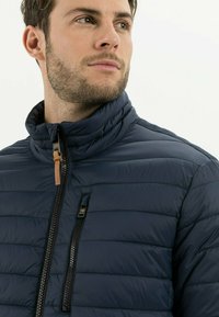 Marineblauer Steppjacke mit hohem Kragen, gestepptem Muster, zwei mit Reißverschluss gesicherten Taschen und Leder-Reißverschlusszugdetail.