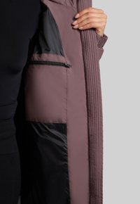 Jachetă quilted purpurie la interior, cu căptușeală neagră solidă și un buzunar cu fermoar. Textură netedă cu detalii în culori contrastante.