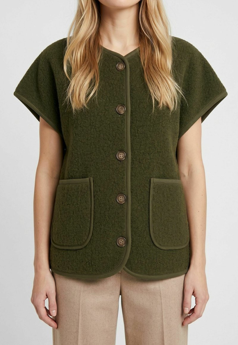 Femme portant un gilet sans manches vert olive boutonné avec des poches avant et un pantalon beige sur un fond uni.