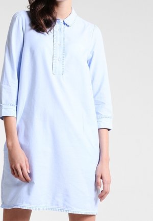 Kobieta ubrana w jasnoniebieską, długorękawową sukienkę do kolan z zapiętym kołnierzykiem na guziki oraz delikatną falbanką na listwie i mankietach.