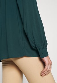 Blouse teal à manches longues avec des poignets froncés, texture lisse et coupe décontractée, associée à un pantalon beige clair à côtes.