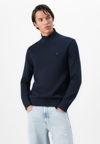 Bărbat îmbrăcat într-un pulover turtleneck albastru marin și blugi deschiși, ușor distresați, stă cu mâinile în buzunare, privind ușor spre lateral.