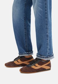 Brūni zamšādas sporta apavi ar melniem akcentiem un oranžām svītrām. Iespējas ar saisināmo dizainu un elastīgu gumijas zoli. Džinsi nedaudz sarullēti pie pēdām.