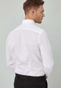 Mand, der står sidelæns iført en hvid langærmet skjorte og sorte dressbukser foran en ensfarvet lys grå baggrund.