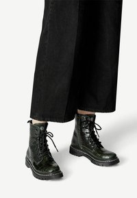 Zwarte combatboots met een glanzend krokodillenpatroon, vetersluiting en een stevige zool. De boots hebben contrasterende stiksels en veterogen.