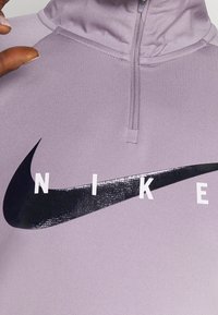 Violetti Nike-poolopaita korkealla kauluksella, jossa puolivetoketju, suuri musta Swoosh-logo ja valkoiset block-kirjaimet. Sileä pinta.