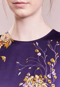Fioletowa tkanina w kwiatowy wzór z żółtymi i różowymi kwiatami, gładka w dotyku, z okrągłym dekoltem. Wykończona delikatnymi konturami kwiatów.
