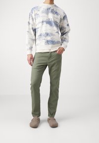 Mies, joka pukeutuu valkoiseen huppariin, jossa on sininen maisemakuva, oliivivihreisiin housuihin, valkoisiin sukkiin ja vaaleanruskeisiin slip-on-kengiin, seisoo valkoista taustaa vasten.