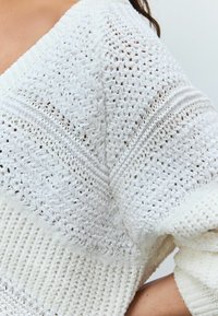 Pull en tricot blanc avec des motifs texturés, présentant une coupe ample et un large décolleté. Le tissu a des motifs de point différents et une finition douce.