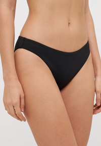 Fekete bikini alsó sima textúrával, magas szabású combnyílásokkal és minimalista dizájnnal. Az anyag rugalmas és a testhez simul.