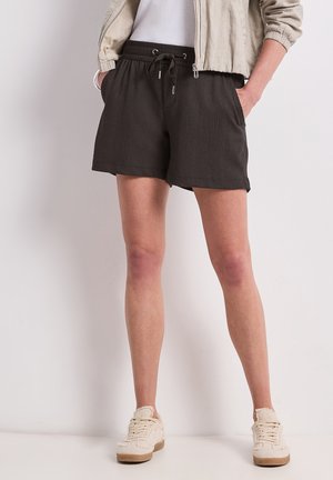 Personne portant un short sombre à cordon, une veste légère, une chemise blanche et des baskets beige, debout les mains dans les poches devant un fond uni.