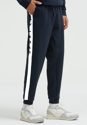 Mann trägt marineblaue Jogginghosen mit weißen Seitenstreifen und schwarzem "SPORT"-Schriftzug, kombiniert mit weißen und grauen Sneakers.