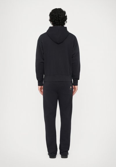 Sudadera negra con capucha y pantalones de chándal a juego, hechos de una tela suave. La sudadera presenta una costura en la parte posterior, mientras que los pantalones tienen un bolsillo trasero con un logo.