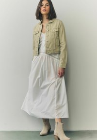 Light khaki denim jacket over a white flowy dress with lace accents, paired with beige ankle boots. Smooth textures and relaxed fit.