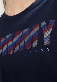 Marineblauses T-Shirt mit dem Aufdruck "TOMMY JEANS" in fetten, gestreiften Buchstaben mit rot, weiß und blauem Muster. Kurze Ärmel, weicher Stoff.