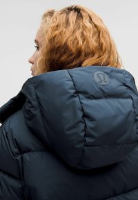 Osoba s kučeravými blond vlasmi, ktorá má na sebe tmavú puffierovú bundu s vyzdvihnutou kapucňou a okrúhlym logom na zadnej strane.