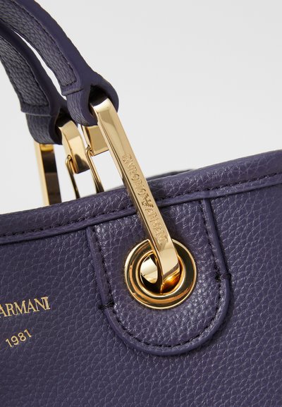 Geantă de piele violet cu accesorii aurii și o etichetă de brand, având mâneri structurate și un finisaj texturat.