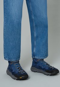Scarpe da trekking blu navy con tomaia testurizzata, lacci grigi e suola in gomma nera con un accento giallo. Indossate con jeans blu a vestibilità ampia.